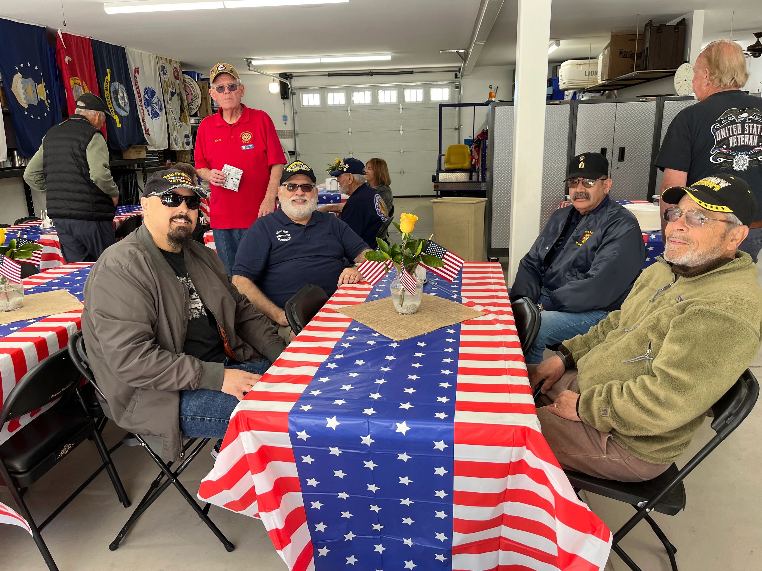 Local veterans seated together at a flag-draped table with service-branch flags on the wall